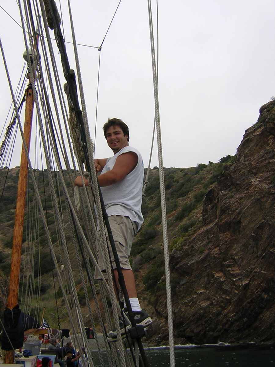 Navarre standing in the Argus rigging, with cliffs behind the ship